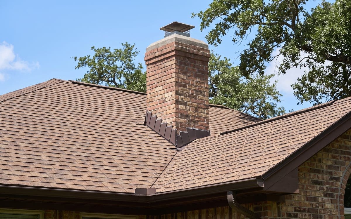 Close view of step flashing and counterflashing where a brown shingle roof meets a brick chimney.