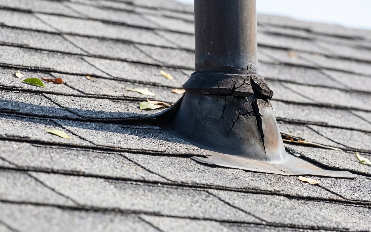 Close view of a cracked rubber collar on a pipe boot on a gray shingle roof, its flange tucked under the upslope shingles.