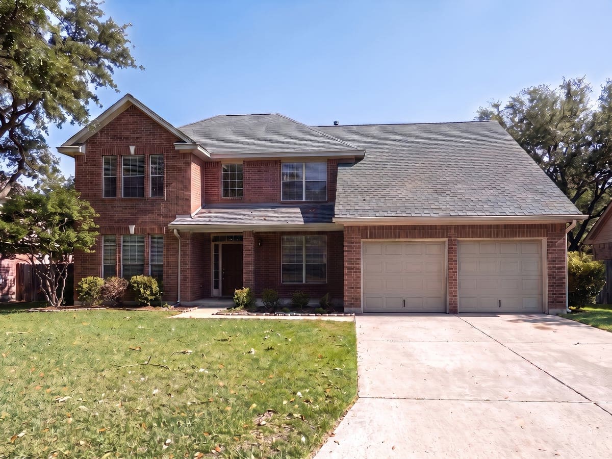 Storm damage roof repair in San Antonio: a brick two-story house with a gray shingle roof on the bright morning after a spring storm, a few small leaves and twigs on the lawn.