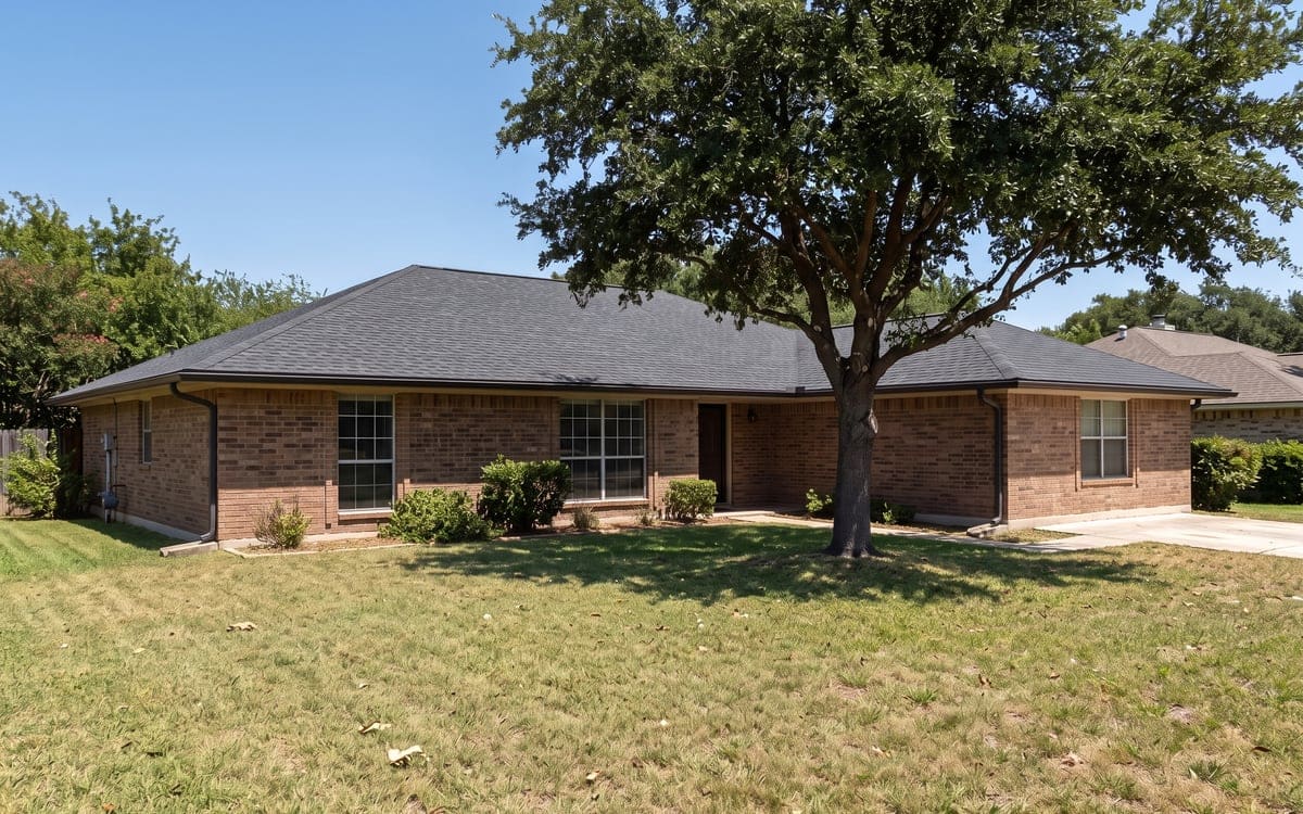 A brick ranch with a charcoal shingle roof under a high summer sun, a live oak casting a short shadow.