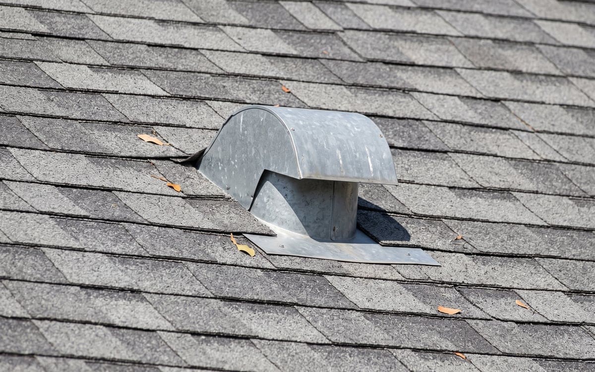 Close view of a metal roof vent cap on a gray shingle roof, its flange under the upslope shingles.