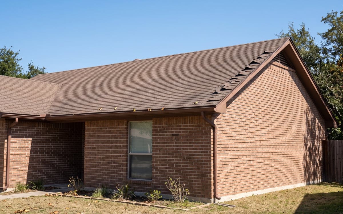 Shingle tabs creased and lifted along the rake edge of a brown shingle roof after high wind, the rest of the roof intact.